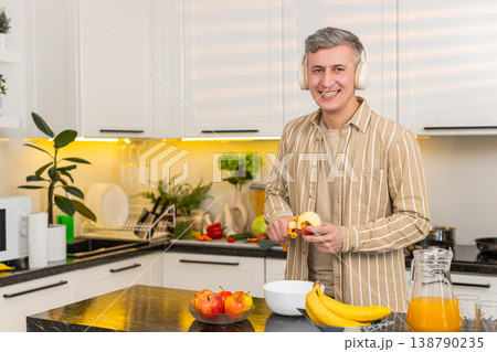 Middle-aged man peeling apple in kitchen with headphones enjoying music during healthy meal routine Middle-aged man peeling apple in kitchen with headphones enjoying music during healthy meal routine 138790235