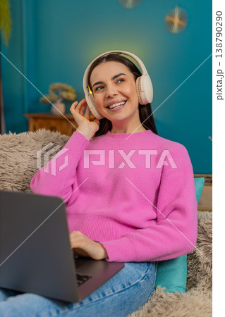 Young woman sitting on sofa wearing headphones listening music while working on laptop with feet up Young woman sitting on sofa wearing headphones listening music while working on laptop with feet up 138790290