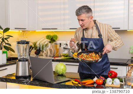 Middle-aged man frying vegetables while reading laptop screen for healthy diet meal in home kitchen 138790710