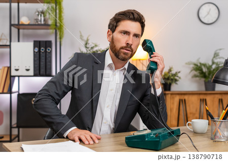 Middle-aged businessman lifts retro wired telephone and makes client call listening closely calmly Middle-aged businessman lifts retro wired telephone and makes client call listening closely calmly 138790718