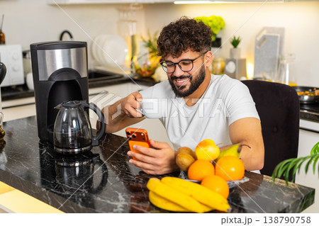 Arabian young adult man drinking fresh hot coffee browsing news on orange smartphone in home kitchen Arabian young adult man drinking fresh hot coffee browsing news on orange smartphone in home kitchen 138790758