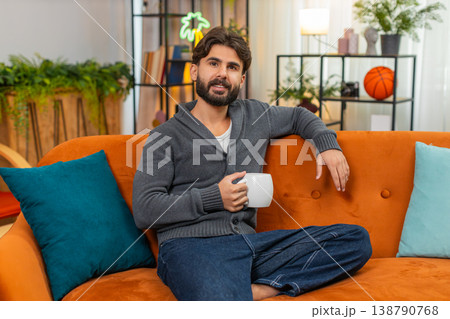 Indian man with hot tea or coffee warms hands gazes out window thoughtful look soft smile calm mood Indian man with hot tea or coffee warms hands gazes out window thoughtful look soft smile calm mood 138790768