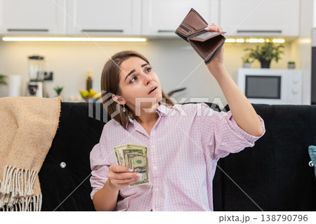 Young woman at home looks inside wallet sees small cash money dollars frowns about poverty debt Young woman at home looks inside wallet sees small cash money dollars frowns about poverty debt 138790796