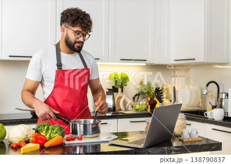 Arabian young adult man tossing hot vegetables in frying pan while watching laptop recipe in kitchen Arabian young adult man tossing hot vegetables in frying pan while watching laptop recipe in kitchen 138790837