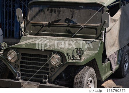 An olive-drab vintage Jeep sits in quiet repose, its caged headlights and weathered canvas top evoking a sense of rugged history against a minimalist urban backdrop. 138790873