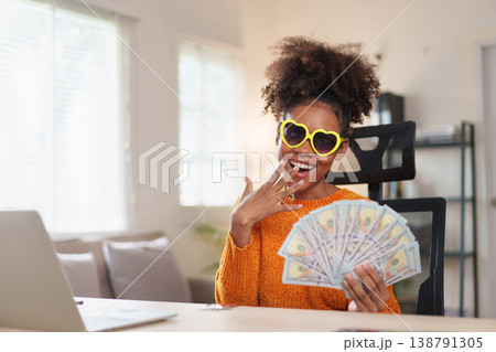 Joyful woman wearing heart-shaped sunglasses, holding a fan of dollar bills, smiling at the camera, expressing financial success and happiness. 138791305