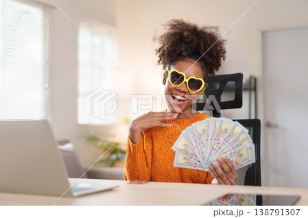 Joyful woman wearing heart-shaped sunglasses, holding a fan of dollar bills, smiling at the camera, expressing financial success and happiness. 138791307