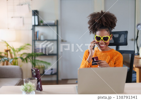 Smiling woman in heart-shaped sunglasses, talking on phone, holding a credit card, sitting at desk with laptop, enjoying online shopping. 138791341