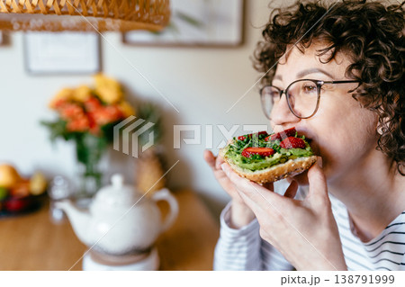 Middle aged woman with curly hair eating avocado toast with strawberries and microgreens in home kitchen morning breakfast healthy food lifestyle natural light close moment Middle aged woman with curly hair eating avocado toast with strawberries and microgreens in home kitchen morning breakfast healthy food lifestyle natural light close moment 138791999