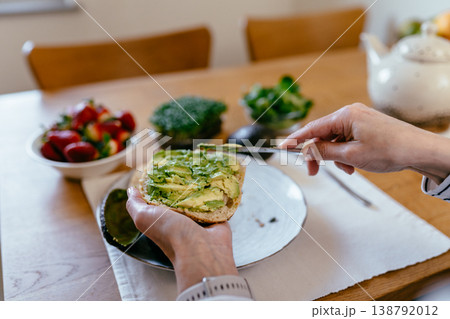 Focused middle aged woman with curly hair spreading avocado on bread with knife plate with strawberries greens microgreens and tea on table home kitchen healthy eating routine Focused middle aged woman with curly hair spreading avocado on bread with knife plate with strawberries greens microgreens and tea on table home kitchen healthy eating routine 138792012