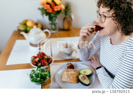 Smiling middle aged woman with curly hair and glasses eating fresh strawberry at breakfast table with avocado bread greens microgreens and tea cozy home kitchen healthy lifestyle routine 138792013
