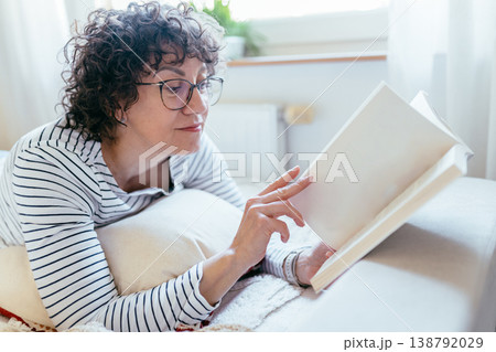 Close up female with glasses lying on bed and reading book. Focused calm mood, cozy indoor setting, quiet leisure, home reading routine, soft daylight, comfort and relaxation. Close up female with glasses lying on bed and reading book. Focused calm mood, cozy indoor setting, quiet leisure, home reading routine, soft daylight, comfort and relaxation. 138792029