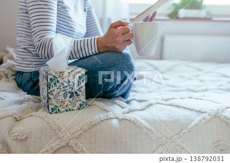 Close up female hands holding mug near book and tissue box on bed. Cozy indoor reading routine, calm home atmosphere, quiet rest, self care, soft textile and daylight. Close up female hands holding mug near book and tissue box on bed. Cozy indoor reading routine, calm home atmosphere, quiet rest, self care, soft textile and daylight. 138792031