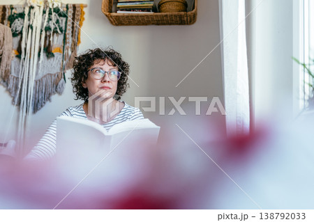 Curly haired female in her 40s with glasses holding book, thoughtful expression, indoor home setting, quiet reading moment, concentration and reflection 138792033