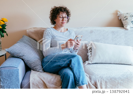 White woman in her 40s sitting on sofa using smartphone, relaxed slight smile, online communication, digital routine, home lifestyle, casual indoor moment, modern technology use 138792041