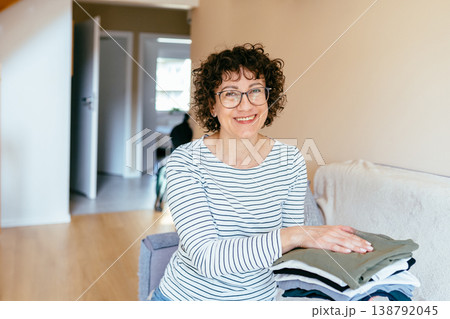 White woman in her 40s smiling at camera while holding folded clothes, satisfied expression, clean organized wardrobe, home routine, neat lifestyle, calm domestic environment 138792045