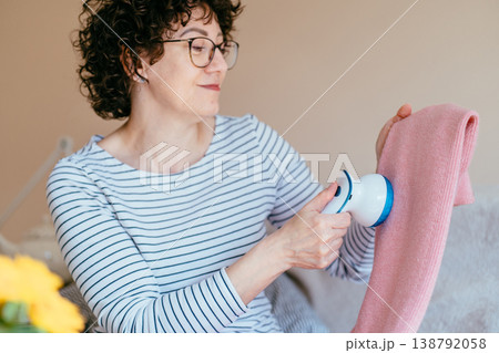 Middle aged woman with glasses gently removing lint from pink knitted sweater with fabric shaver. Calm focused mood, clothing care routine, cozy home environment, slow living concept. Middle aged woman with glasses gently removing lint from pink knitted sweater with fabric shaver. Calm focused mood, clothing care routine, cozy home environment, slow living concept. 138792058