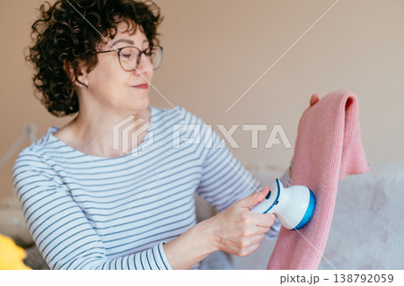 Middle aged woman with glasses holding pink sweater and using fabric shaver. Soft focus, relaxed mood, clothing care routine, gentle maintenance, cozy home lifestyle, everyday comfort. Middle aged woman with glasses holding pink sweater and using fabric shaver. Soft focus, relaxed mood, clothing care routine, gentle maintenance, cozy home lifestyle, everyday comfort. 138792059