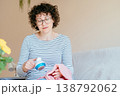 Middle aged woman with curly hair and glasses gently cleaning pink sweater with fabric shaver while sitting on sofa. Calm expression, home routine, clothing care, slow living, cozy domestic moment. 138792062