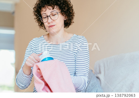 Middle aged woman with glasses carefully cleaning pink knitwear using fabric shaver. Concentrated expression, hands working on textile, clothing maintenance, pilling removal, everyday home routine. Middle aged woman with glasses carefully cleaning pink knitwear using fabric shaver. Concentrated expression, hands working on textile, clothing maintenance, pilling removal, everyday home routine. 138792063