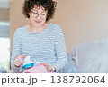 Middle aged woman with curly hair and glasses using fabric shaver on pink knitted sweater while sitting on sofa. Focused expression, clothing care, lint removal, home textile maintenance routine. 138792064