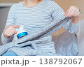 Unrecognizable woman in striped shirt stretching gray knitted blanket and removing lint with fabric shaver while sitting on sofa. Clothing care, pilling removal, textile maintenance at home routine. 138792067