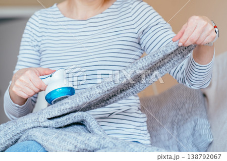 Unrecognizable woman in striped shirt stretching gray knitted blanket and removing lint with fabric shaver while sitting on sofa. Clothing care, pilling removal, textile maintenance at home routine. 138792067