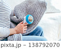 Cropped woman uses electric fabric shaver on gray knitted fabric while resting on sofa at home. Detail of hands, textile care, sweater cleaning, pilling removal, domestic routine, cozy maintenance. 138792070