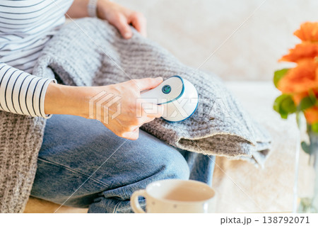 Close up of woman holding fabric shaver above gray knitted clothing during home care routine. unrecognizable person, soft textile, garment upkeep, lint removal, domestic maintenance. 138792071