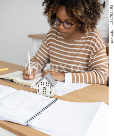 female architect designer is working, taking notes in notebook is mock up of house on table. 138792392