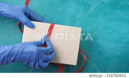 Pandemic present. Gift wrapping. Hands in protective gloves opening box untying red bow on blue textured free space background. Pandemic present. Gift wrapping. Hands in protective gloves opening box untying red bow on blue textured free space background. 138792641