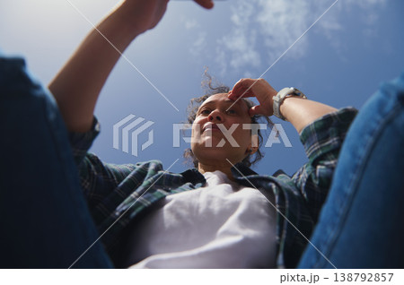 Low angle view of woman using smartphone outdoors against bright blue sky in sunny park environment Low angle view of woman using smartphone outdoors against bright blue sky in sunny park environment 138792857