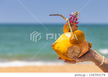 Fresh tropical beverage in a vibrant yellow coconut, garnished with flowers, held by a hand against a sunny beach and ocean background. Sri Lanka island in Asia. Fresh tropical beverage in a vibrant yellow coconut, garnished with flowers, held by a hand against a sunny beach and ocean background. Sri Lanka island in Asia. 138793179
