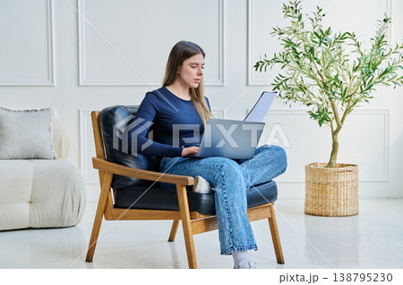 Portrait of young woman sitting on chair with laptop, in home interior Portrait of young woman sitting on chair with laptop, in home interior 138795230