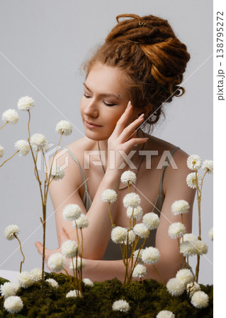 Young Caucasian redhead woman touching face among dried white flowers on moss, white wall Young Caucasian redhead woman touching face among dried white flowers on moss, white wall 138795272