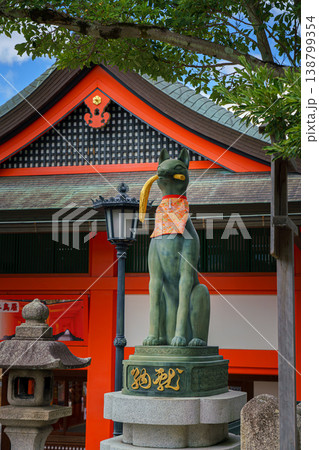 Kyoto, Japan - Sep 23 2024, Close-up view of the stone statue of kitsune, a Japanese fox holding a golden object in its mouth, with red facade of Fushimi-Inari Temple in background, Kyoto, Japan 138799354