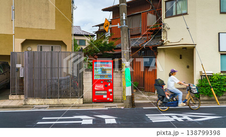 Tokyo, Japan - Sep 30 2024, panoramic view of street in a residential area with private houses, there is a red vending machine with drinks on side of road, a woman rides by on a bicycle, Tokyo, Japan Tokyo, Japan - Sep 30 2024, panoramic view of street in a residential area with private houses, there is a red vending machine with drinks on side of road, a woman rides by on a bicycle, Tokyo, Japan 138799398