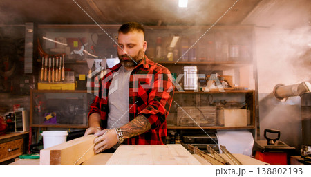 Joinery process. Stacking wood. Male carpenter checking plank quality holding timber on workbench in smoky workshop with equipment. 138802193