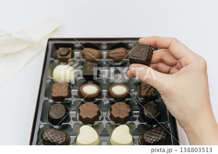 白い背景に置かれたチョコレートボックスから、1粒を指先で摘み上げる女性の手元 白い背景に置かれたチョコレートボックスから、1粒を指先で摘み上げる女性の手元 138803513