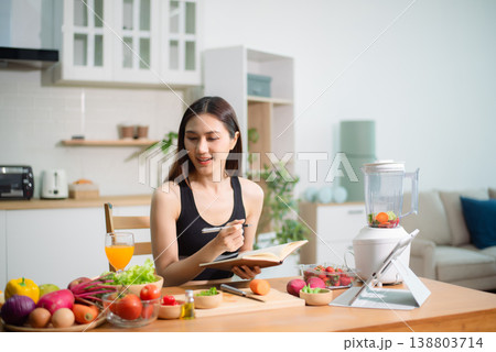 Asian woman reading recipe book while preparing fresh salad in home kitchen. Clean eating, nutrition planning, meal prep and healthy lifestyle concept. 138803714
