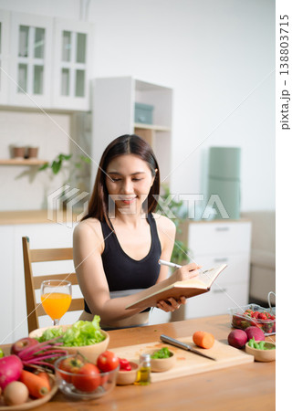 Asian woman reading recipe book while preparing fresh salad in home kitchen. Clean eating, nutrition planning, meal prep and healthy lifestyle concept. Asian woman reading recipe book while preparing fresh salad in home kitchen. Clean eating, nutrition planning, meal prep and healthy lifestyle concept. 138803715