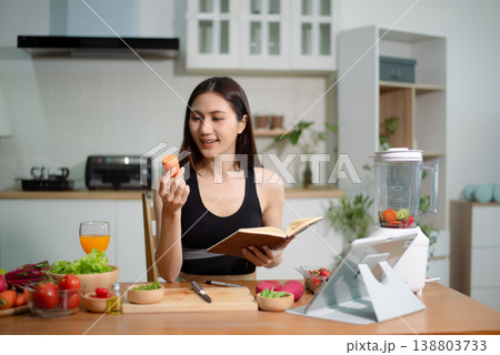 Asian woman reading recipe book while preparing fresh salad in home kitchen. Clean eating, nutrition planning, meal prep and healthy lifestyle concept. 138803733