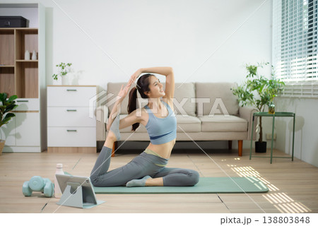Asian woman doing yoga stretching on mat at home with online workout. Home fitness, flexibility training, wellness lifestyle, self care, indoor exercise. 138803848