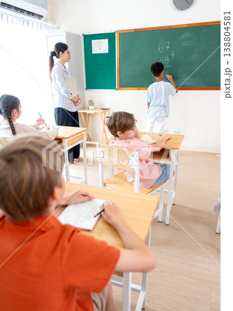 Vertical image of the teacher tell African American boy try to write the answer for calculation question on blackboard in front of classroom. Vertical image of the teacher tell African American boy try to write the answer for calculation question on blackboard in front of classroom. 138804581