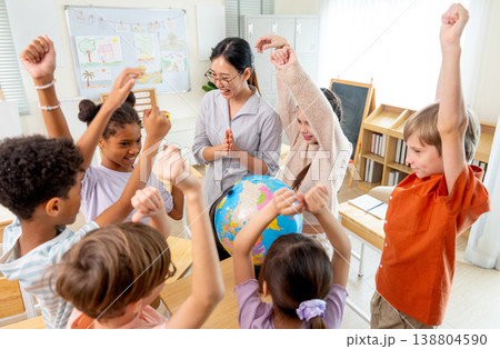 High view of teacher give the question about country on earth and tell the kids to answer and the children raise their hands to compete for answer using model globe. High view of teacher give the question about country on earth and tell the kids to answer and the children raise their hands to compete for answer using model globe. 138804590
