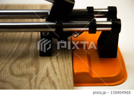 Close-up side view of an orange mechanical tile cutter, which fixed ceramic tiles of wooden colors, above it is a cutting disc on a white background. The concept of renovation, tiling, construction 138804988