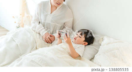 ベッドルームで男の子をスマホを使って寝かしつける母親 ベッドルームで男の子をスマホを使って寝かしつける母親 138805535