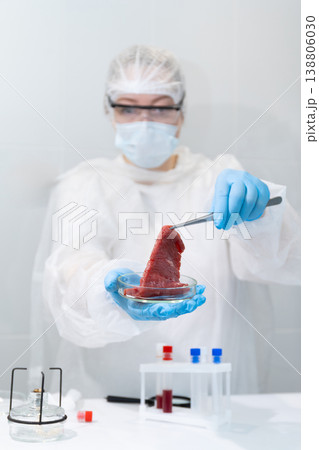 Woman scientist in protective gear handling lab-grown meat sample with tongs in petri dish. Artificial meat cultivation in biotechnology laboratory. Sustainable protein production and innovative food 138806030