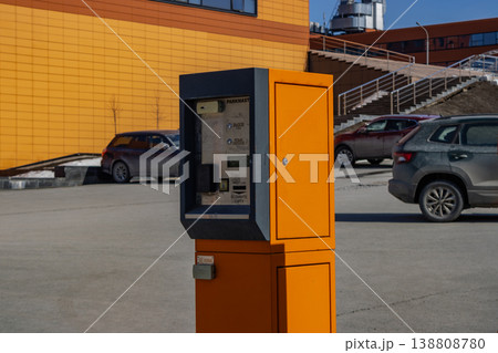 A Bright Orange Parking Meter Standing Against a Modern Urban Background with Cars Parked Nearby and a Clear Blue Sky Above A Bright Orange Parking Meter Standing Against a Modern Urban Background with Cars Parked Nearby and a Clear Blue Sky Above 138808780