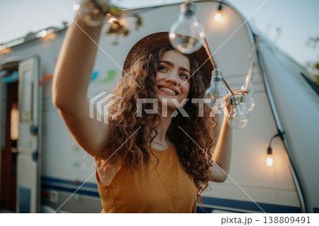 Young beautiful woman is on a camping trip in nature, putting lights on tree, preparing for evening. Young beautiful woman is on a camping trip in nature, putting lights on tree, preparing for evening. 138809491